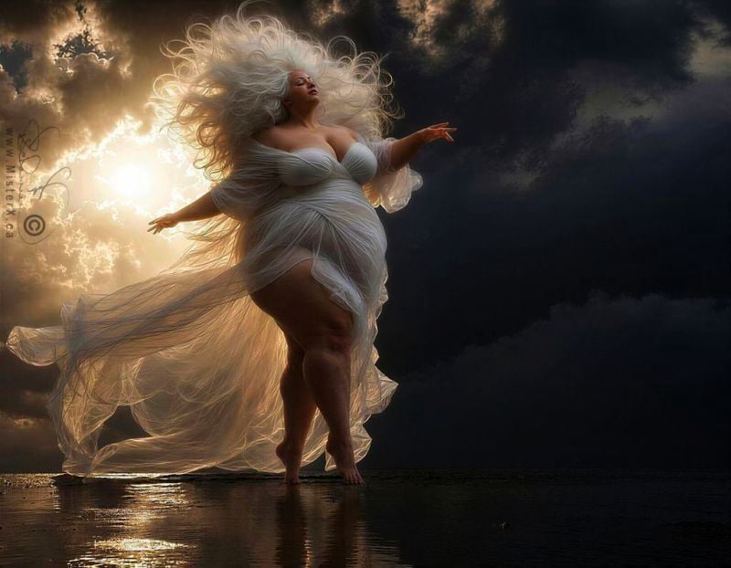 A low-angle shot captures a plus-size woman with flowing white hair and a white, translucent flowing gown standing on the shore at dawn or dusk. The right side of the image features a dramatic, dark, cloudy sky, while the left side is bathed in a golden light from behind the clouds. Her head is tilted back, eyes closed, and her arms are outstretched. Her white gown billows around her feet, revealing glimpses of her legs, which are reflected in the wet sand.