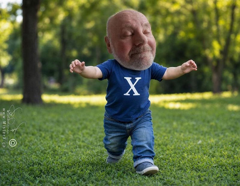 A highly detailed full body shot of a toddler with the head of a bald elderly man with a white beard, wearing a navy blue plain t-shirt with a white 'X' and light blue jeans, walking on green grass in a park. His eyes are closed and he has a subtle smile, with his arms outstretched to the sides. The background features lush green foliage and trees, blurred to create a sense of depth and focus on the subject. The lighting is bright and natural, suggesting an outdoor setting on a sunny day.