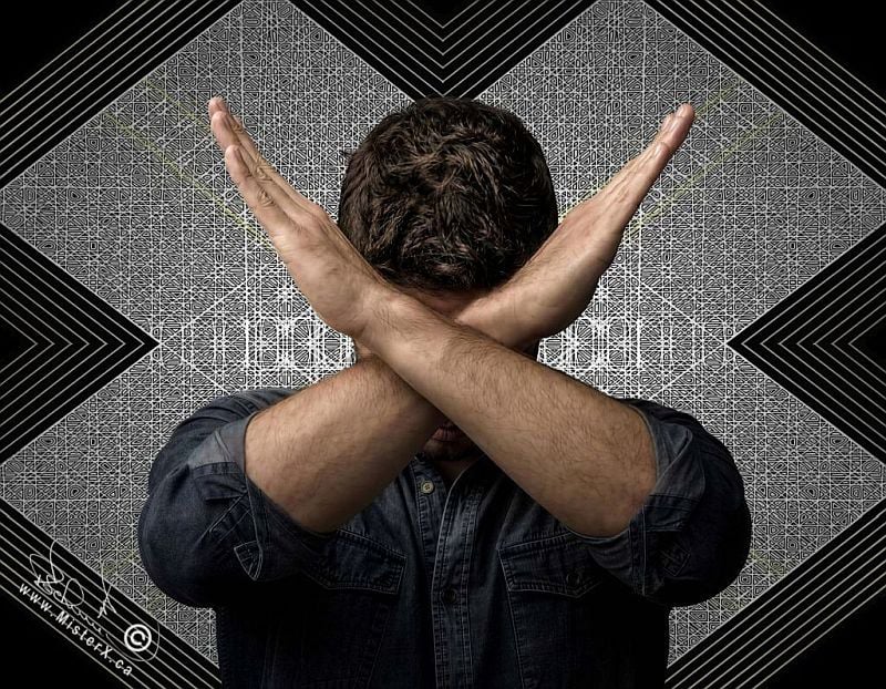 A man is seen standing in front of a detailed letter "X". His face is covered as he crosses his arms in front of it in an "X" shape.